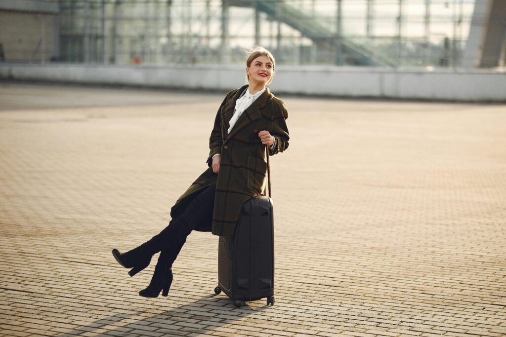 Smiling woman in a plaid coat sits on luggage at an airport terminal, enjoying a winter day.